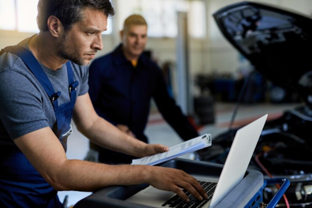 auto repairman working on a computer while doing car diagnostic with his coworker in a workshop to deal with challenges in tеlеmatics